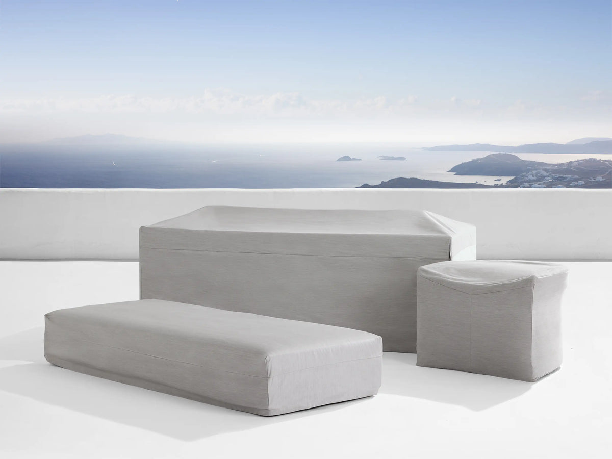 Grey outdoor chair cover on a patio, tailored fit over a chair. Smooth fabric with front slit, set against a scenic ocean view background.