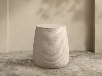 Eden Outdoor End Table. Light tan, round concrete table with distinct horizontal ribbed sides, on a stone patio.