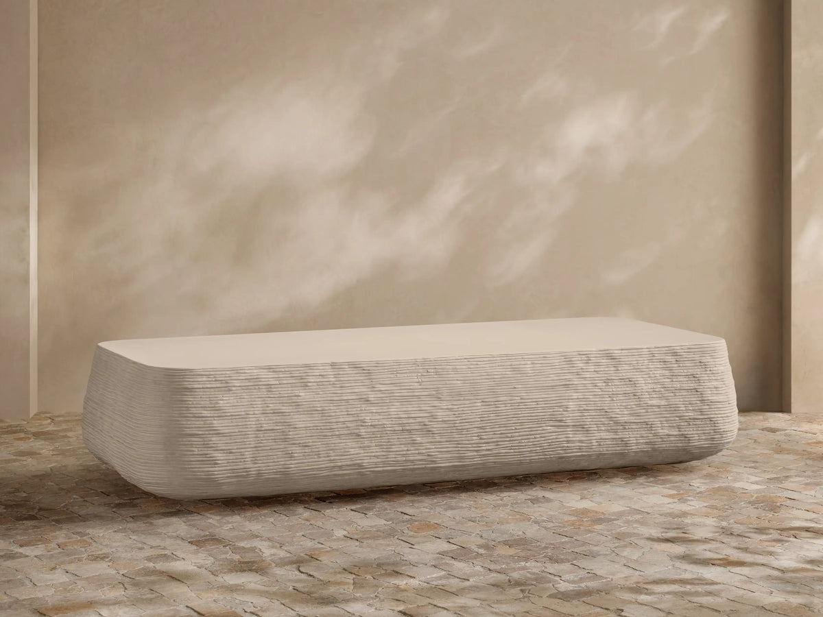 Eden Outdoor Coffee Table, off-white, rectangular with rounded edges, horizontal ribbed texture on sides, against tan wall and stone floor.