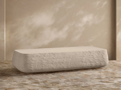 Eden Outdoor Coffee Table, off-white, rectangular with rounded edges, horizontal ribbed texture on sides, against tan wall and stone floor.