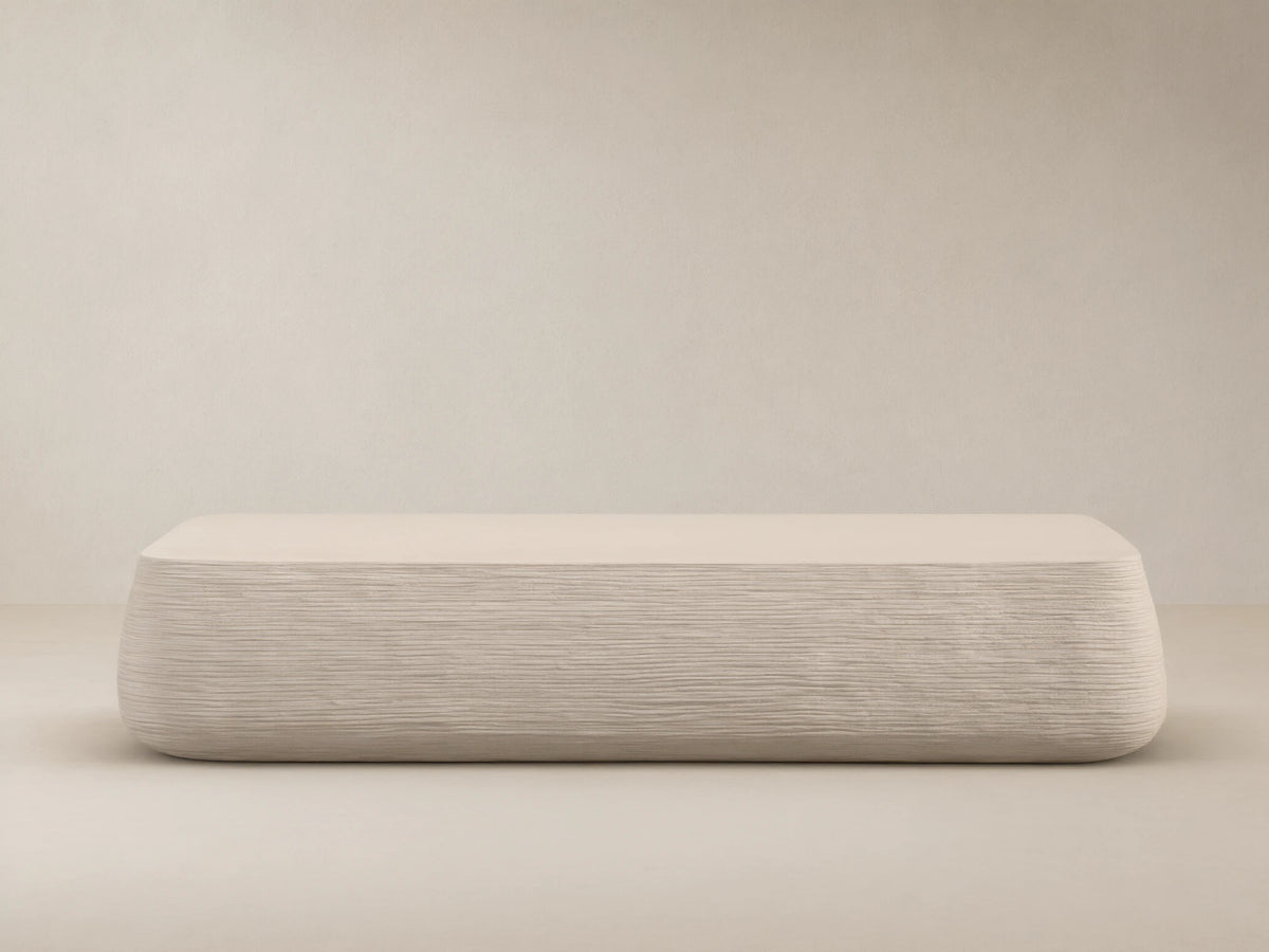 Eden Outdoor Coffee Table, off-white, rectangular with rounded edges, horizontal ribbed texture on sides, against tan wall and stone floor.