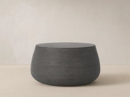 Dark grey Eden Outdoor Round Coffee Table with textured ribbed sides and smooth top, on a light brown tiled patio.