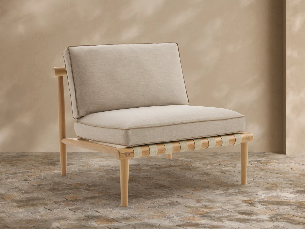 Elio Outdoor Lounge Chair with natural teak frame, light beige piped cushions, and woven seat support on a patio.