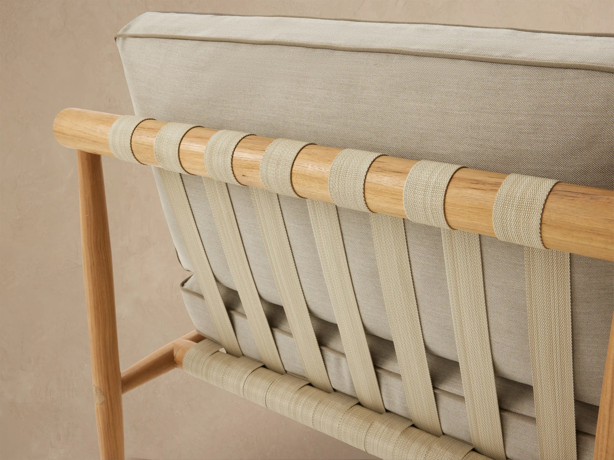 Elio Outdoor Lounge Chair, front view. Light teak wood frame with woven beige straps, thick cream cushions with brown piping on a tiled patio.