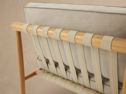 Elio Outdoor Lounge Chair, front view. Light teak wood frame with woven beige straps, thick cream cushions with brown piping on a tiled patio.