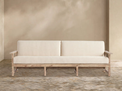 Lofty, light beige Emmons Outdoor Replacement Cushions on a wooden sofa frame, against a tan wall and stone patio.