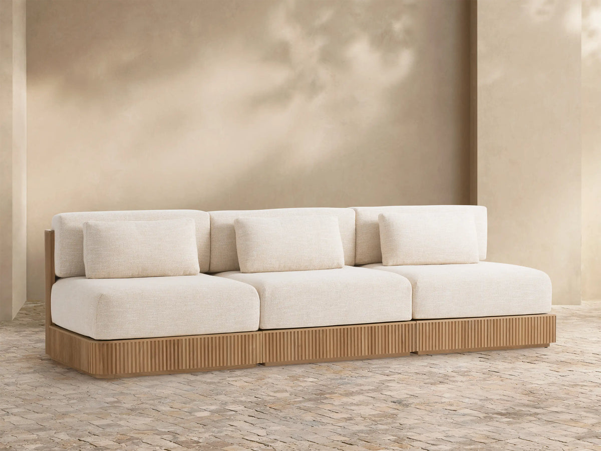 Beaumont Outdoor Teak Three-Piece Modular Sofa, natural slatted teak base, light cream cushions, against a warm beige wall.