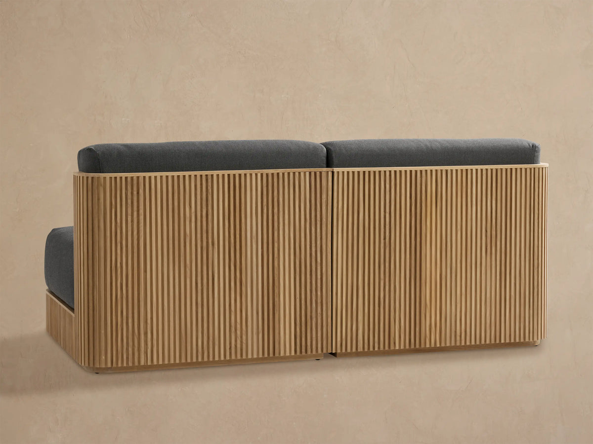 Angled view of Beaumont Outdoor Teak Modular Sofa corner section. Light ribbed teak frame, dark gray seat and back cushions.
