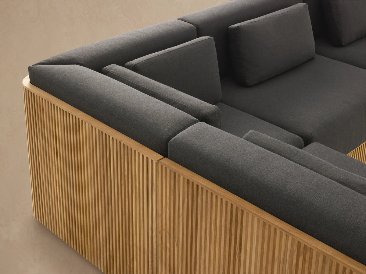 Beaumont Outdoor Teak Six-Piece Sectional with Chaise: L-shaped, natural slatted teak base, dark grey cushions, on a light patio.