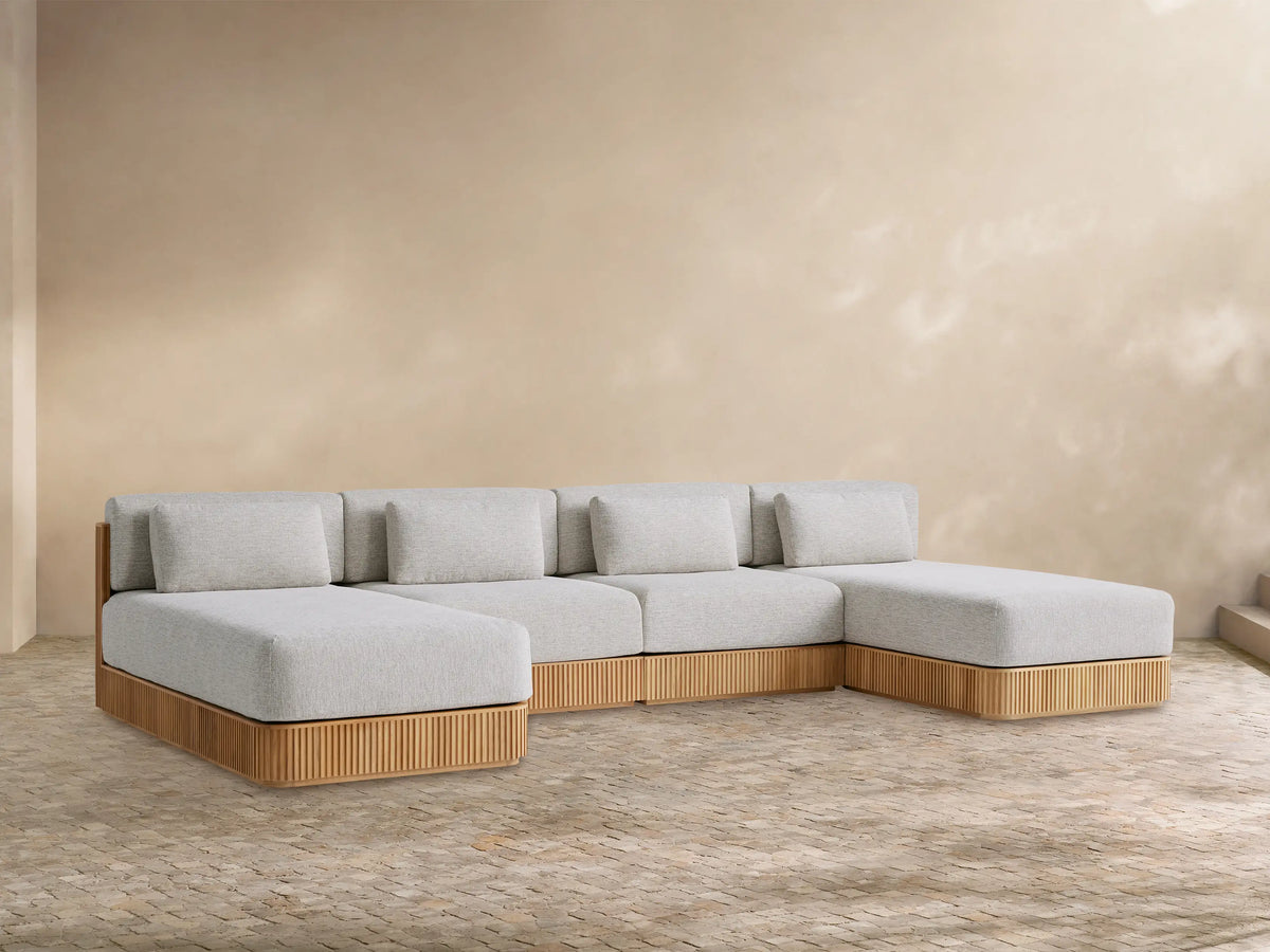 Beaumont Outdoor Four-Piece Double Chaise Sectional, light grey cushions, natural teak slatted base on textured floor, beige wall.