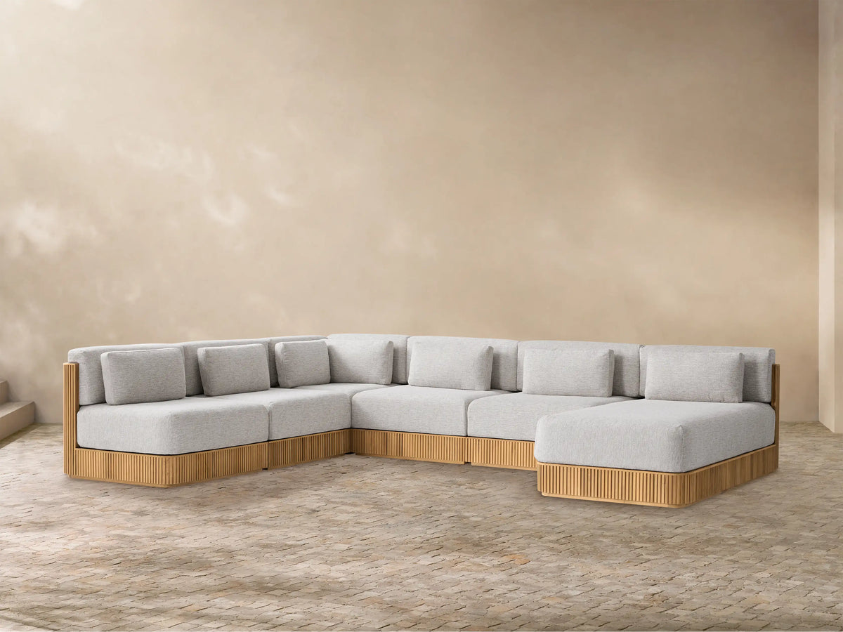 L-shaped Beaumont Outdoor Teak Sectional with light gray cushions, natural slatted teak base, chaise on right, on a textured floor.