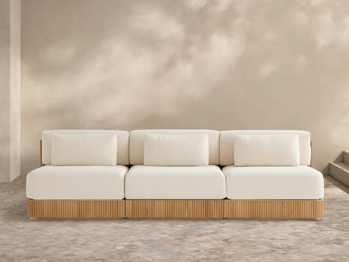 Beaumont Outdoor Teak Three-Piece Modular Sofa with light cream cushions & ribbed teak base on a tiled patio, beige wall behind.