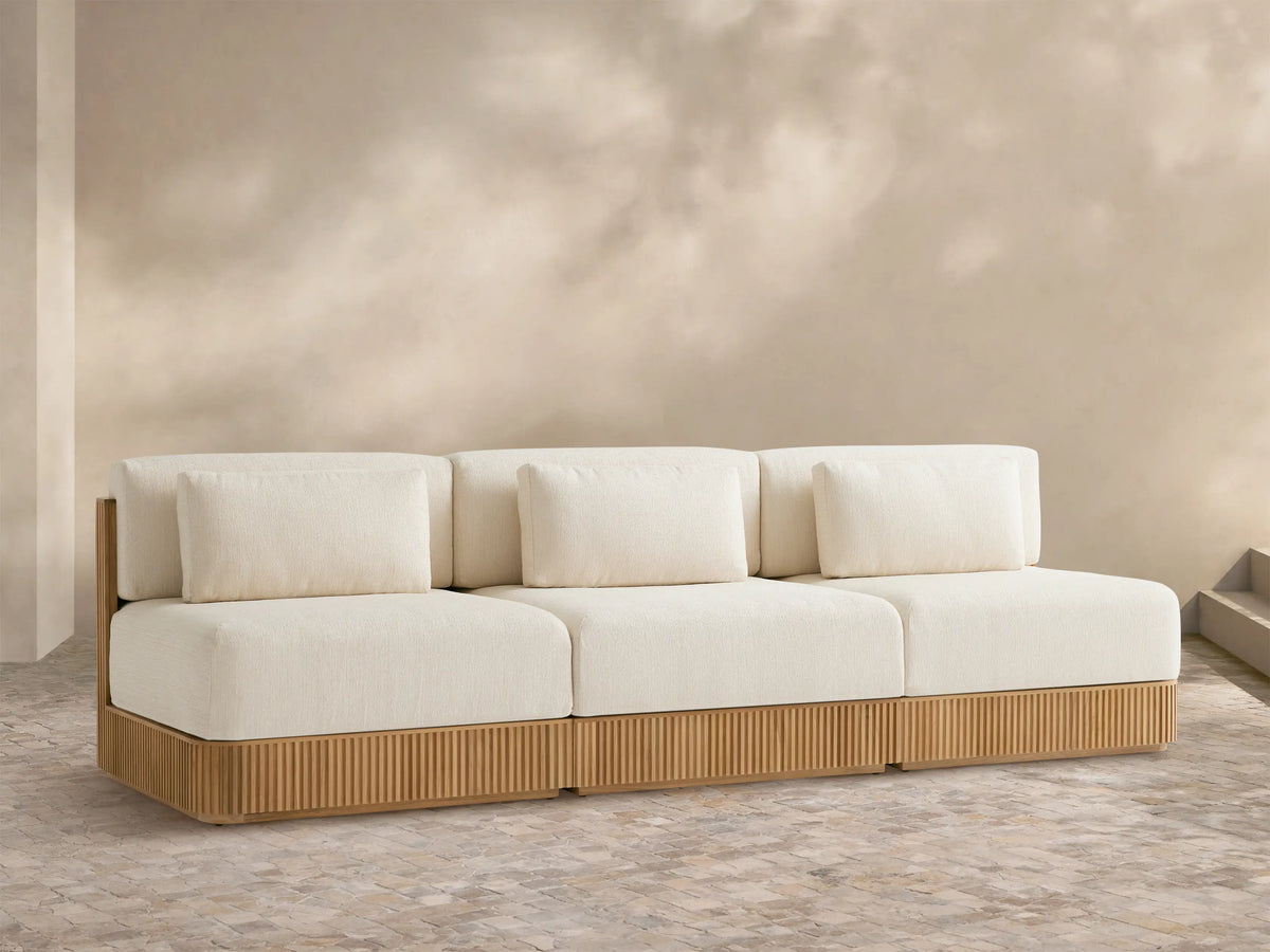 Beaumont Outdoor Teak Three-Piece Modular Sofa with light cream cushions & ribbed teak base on a tiled patio, beige wall behind.