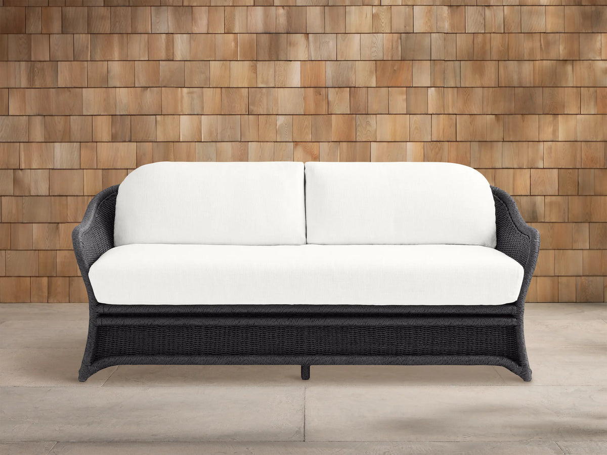 Fowler Outdoor Sofa: Dark gray woven frame with two plush white cushions, set on a concrete patio in front of a wood shingle wall.