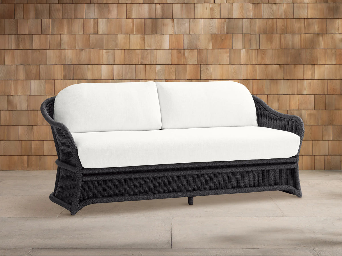 Fowler Outdoor Sofa: Dark gray woven frame with two plush white cushions, set on a concrete patio in front of a wood shingle wall.