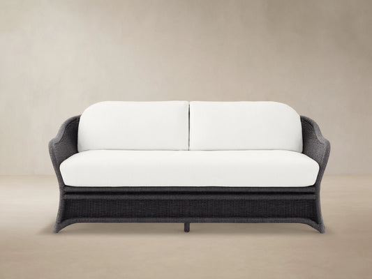Fowler Outdoor Sofa: Dark gray woven frame with two plush white cushions, set on a concrete patio in front of a wood shingle wall.