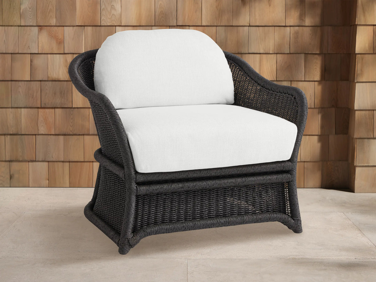Fowler Outdoor Lounge Chair with dark gray handwoven wicker frame & white cushions, angled against a wood shingle wall & patio.