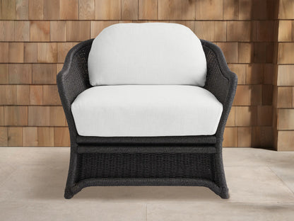 Fowler Outdoor Lounge Chair with dark gray handwoven wicker frame & white cushions, angled against a wood shingle wall & patio.