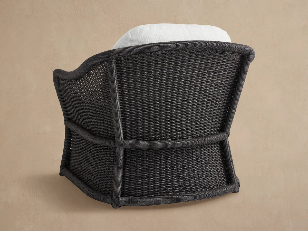 Close-up of Fowler Outdoor Lounge Chair, angled. Black woven wicker frame, thick white seat and back cushions, against beige wall.