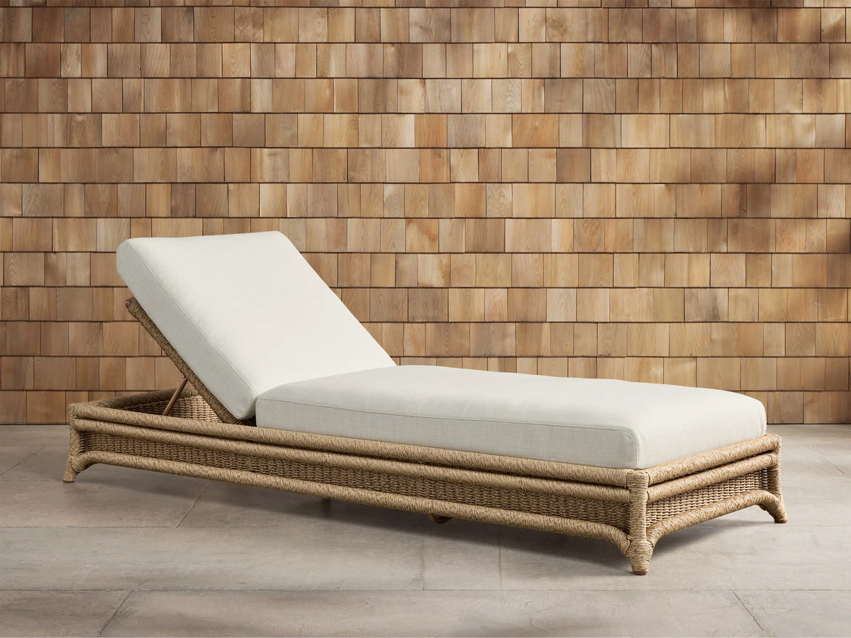 Fowler Outdoor Chaise with inclined backrest, light cushions, and natural woven wicker frame on a patio by a shingle wall.