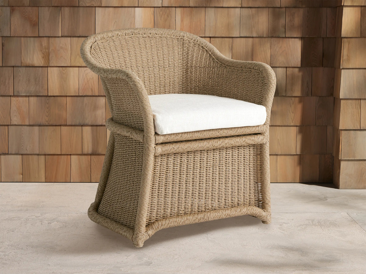 Fowler Outdoor Dining Chair, light tan woven wicker with curved back and arms, off-white cushion, on stone floor against wood wall.