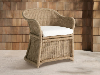 Fowler Outdoor Dining Chair, light tan woven wicker with curved back and arms, off-white cushion, on stone floor against wood wall.