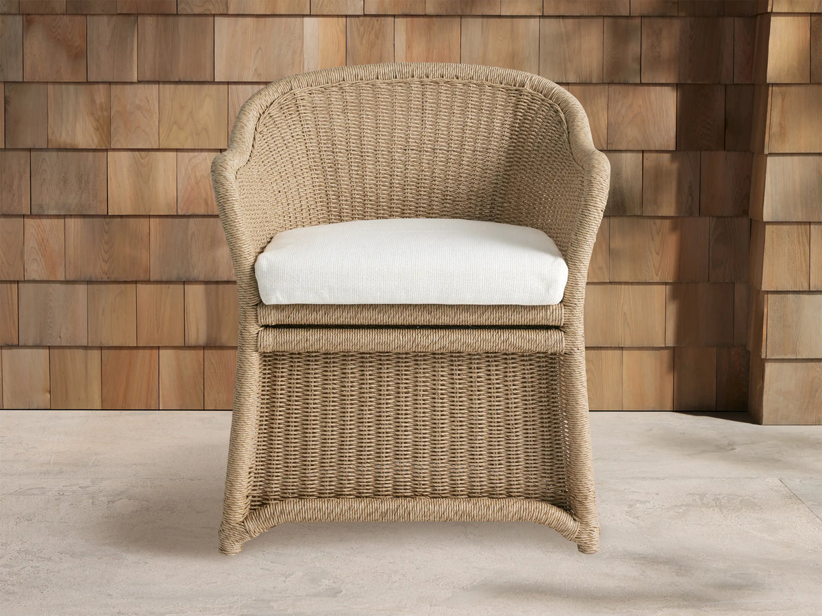 Fowler Outdoor Dining Chair, light tan woven wicker with curved back and arms, off-white cushion, on stone floor against wood wall.