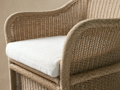 Fowler Outdoor Dining Chair: natural woven wicker with rounded back & arms, white cushion, on a patio against wood wall.