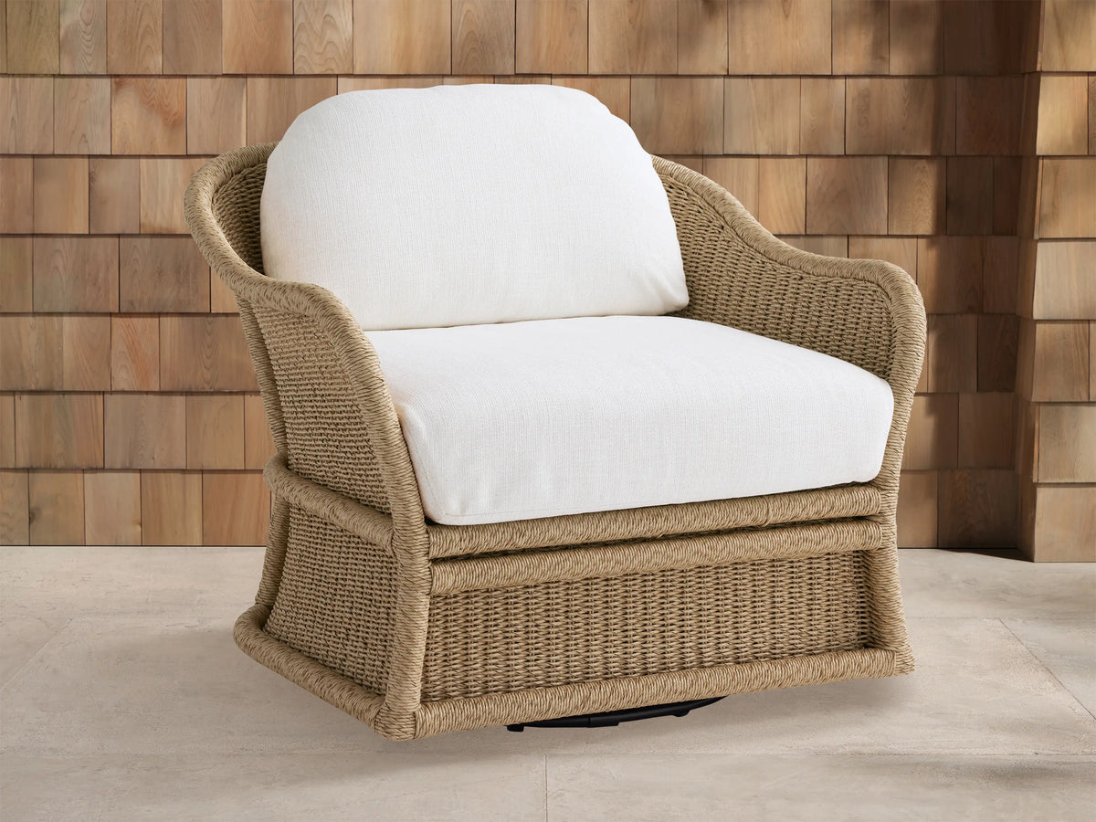 Fowler Outdoor Glider, light brown woven wicker chair with white cushions, angled on light patio tiles against a wood-paneled wall.
