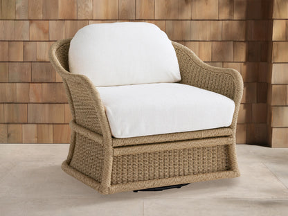 Fowler Outdoor Glider, light brown woven wicker chair with white cushions, angled on light patio tiles against a wood-paneled wall.