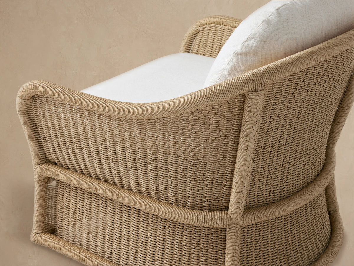 Fowler Outdoor Glider: Tan handwoven all-weather wicker chair with curved arms, plush white cushions, against a wood shingle wall on a patio.