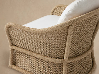 Fowler Outdoor Glider: Tan handwoven all-weather wicker chair with curved arms, plush white cushions, against a wood shingle wall on a patio.