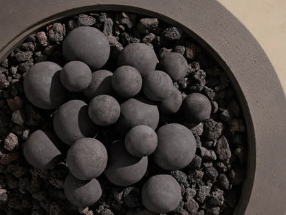 Black outdoor fire pit spheres, various sizes, on dark lava rocks in a textured grey concrete fire pit.
