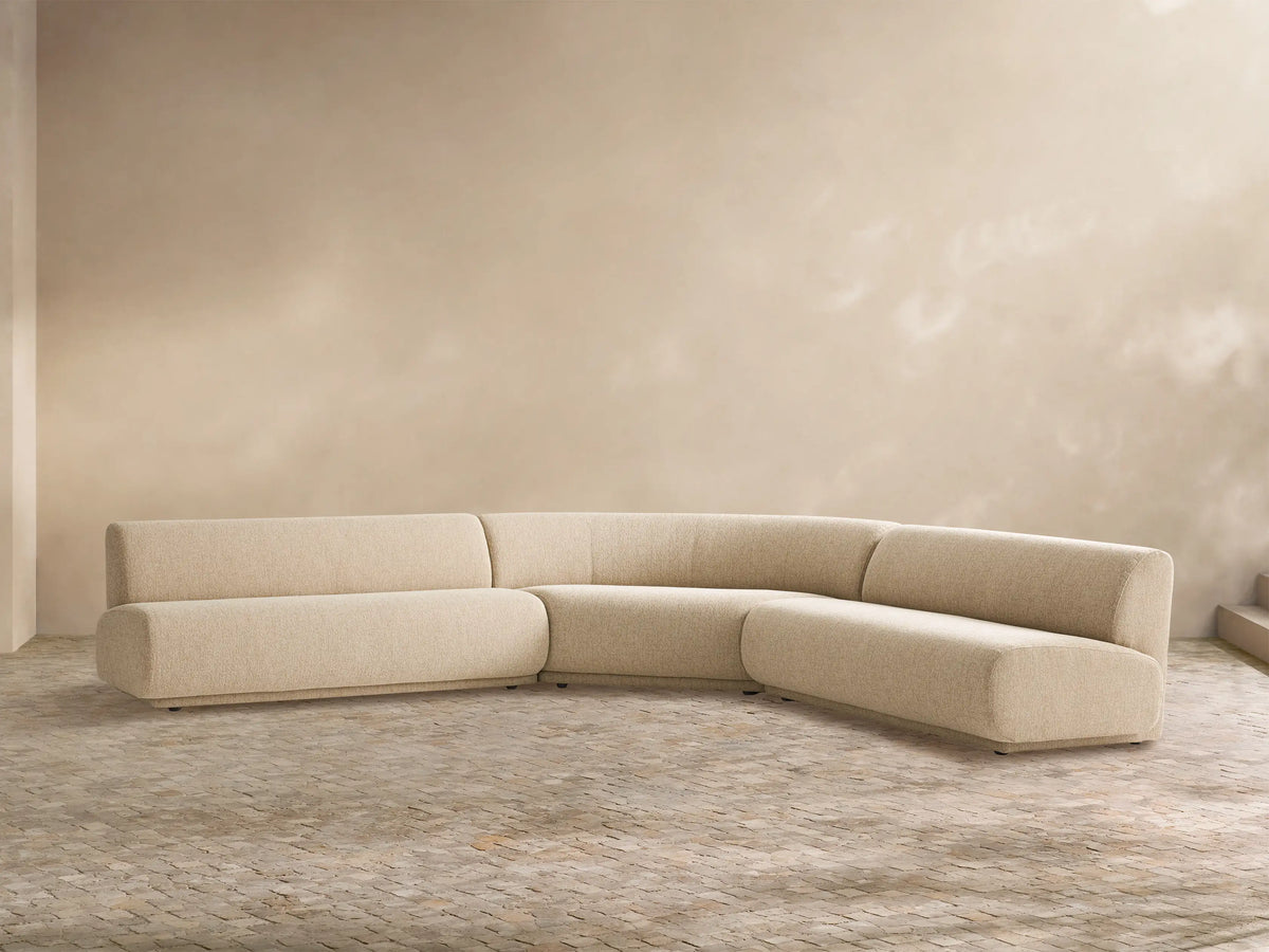 Gianni Outdoor Sectional: Sand-colored, L-shaped sofa with curved armless section, on a light tiled floor against a neutral wall.