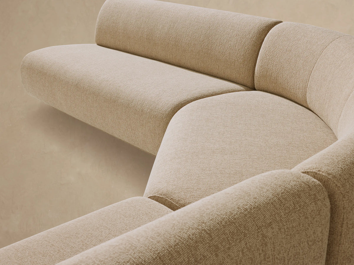 Light beige Gianni Outdoor Sectional, curved and modular, sits on a stone-tiled patio in front of a warm, sandy wall.