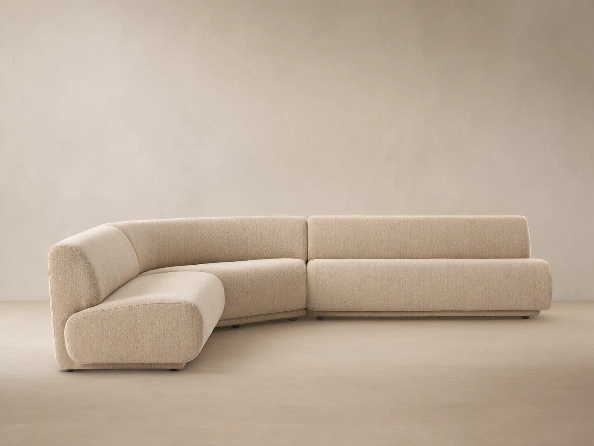 Gianni Outdoor Sectional: Sand-colored, L-shaped sofa with curved armless section, on a light tiled floor against a neutral wall.
