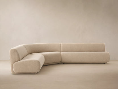 Gianni Outdoor Sectional: Sand-colored, L-shaped sofa with curved armless section, on a light tiled floor against a neutral wall.