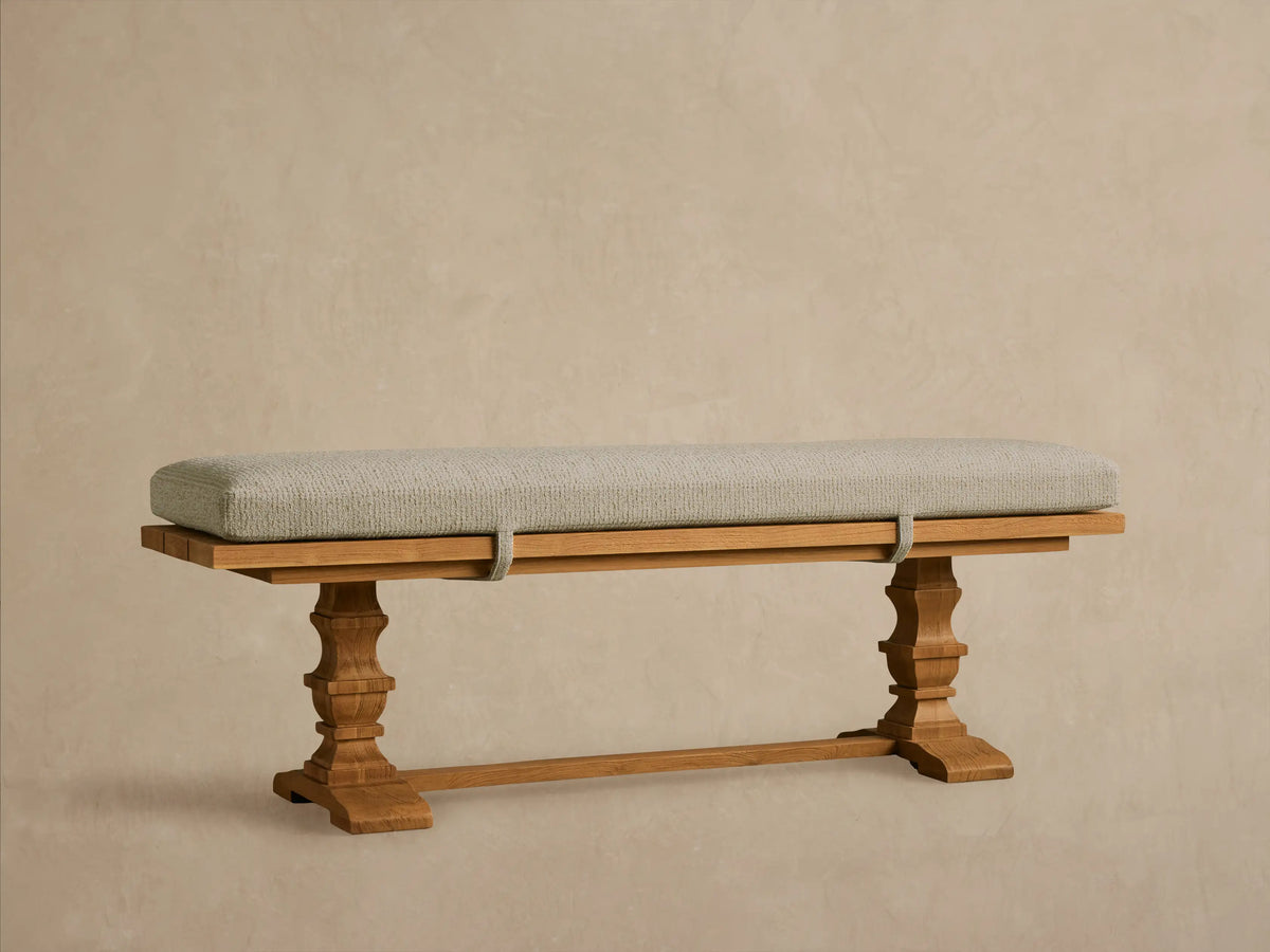 Hamptons Outdoor Dining Bench: light brown teak wood with ornate legs, long light green cushion, against a wood shingle wall.