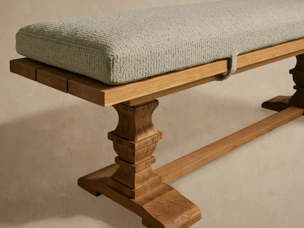 Hamptons outdoor dining bench, light brown teak wood base with ornate carved legs, light grey cushion secured by straps.