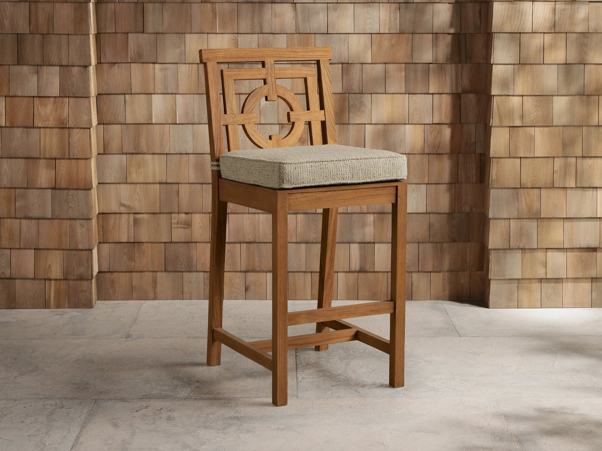 Weathered fawn teak Hamptons Outdoor Barstool with geometric backrest, light beige cushion, against wood shingle wall.