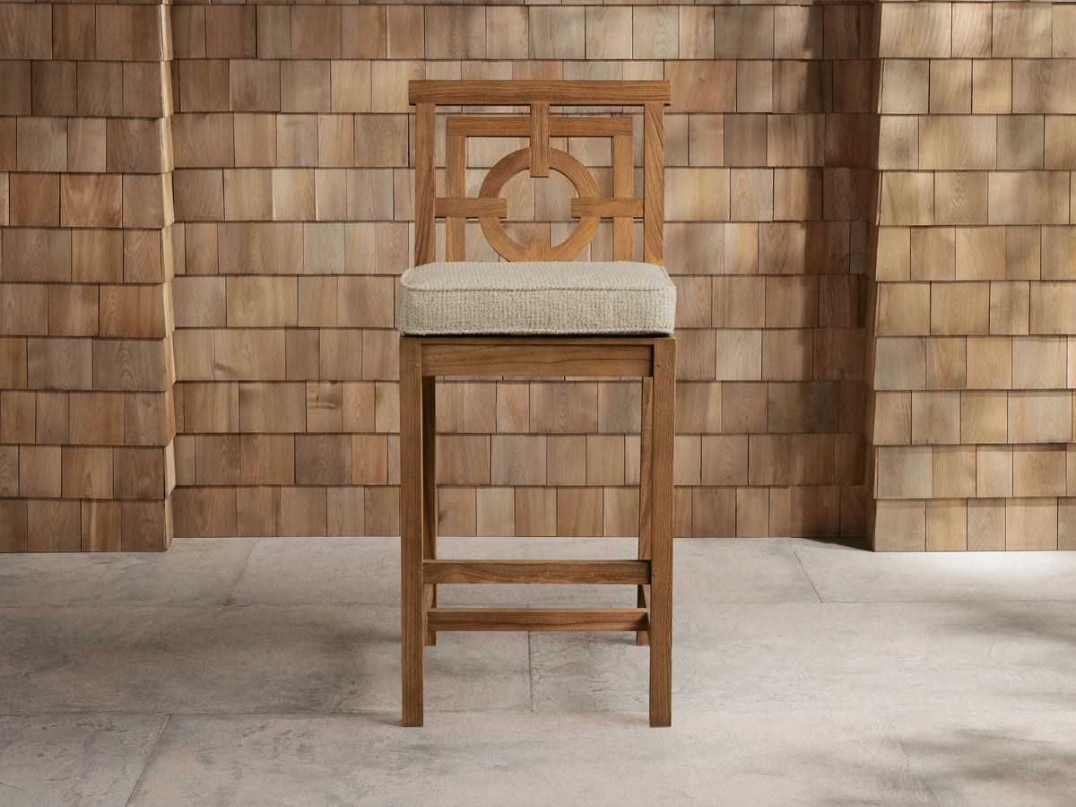 Weathered fawn teak Hamptons Outdoor Barstool with geometric backrest, light beige cushion, against wood shingle wall.