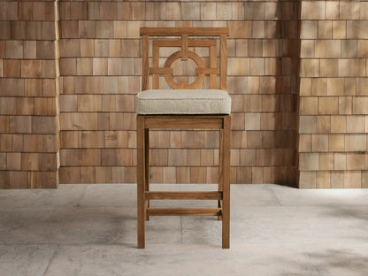 Weathered fawn teak Hamptons Outdoor Barstool with geometric backrest, light beige cushion, against wood shingle wall.