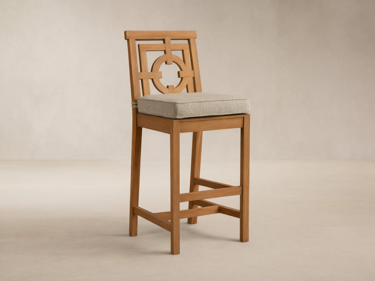 Weathered fawn teak Hamptons Outdoor Barstool with geometric backrest, light beige cushion, against wood shingle wall.