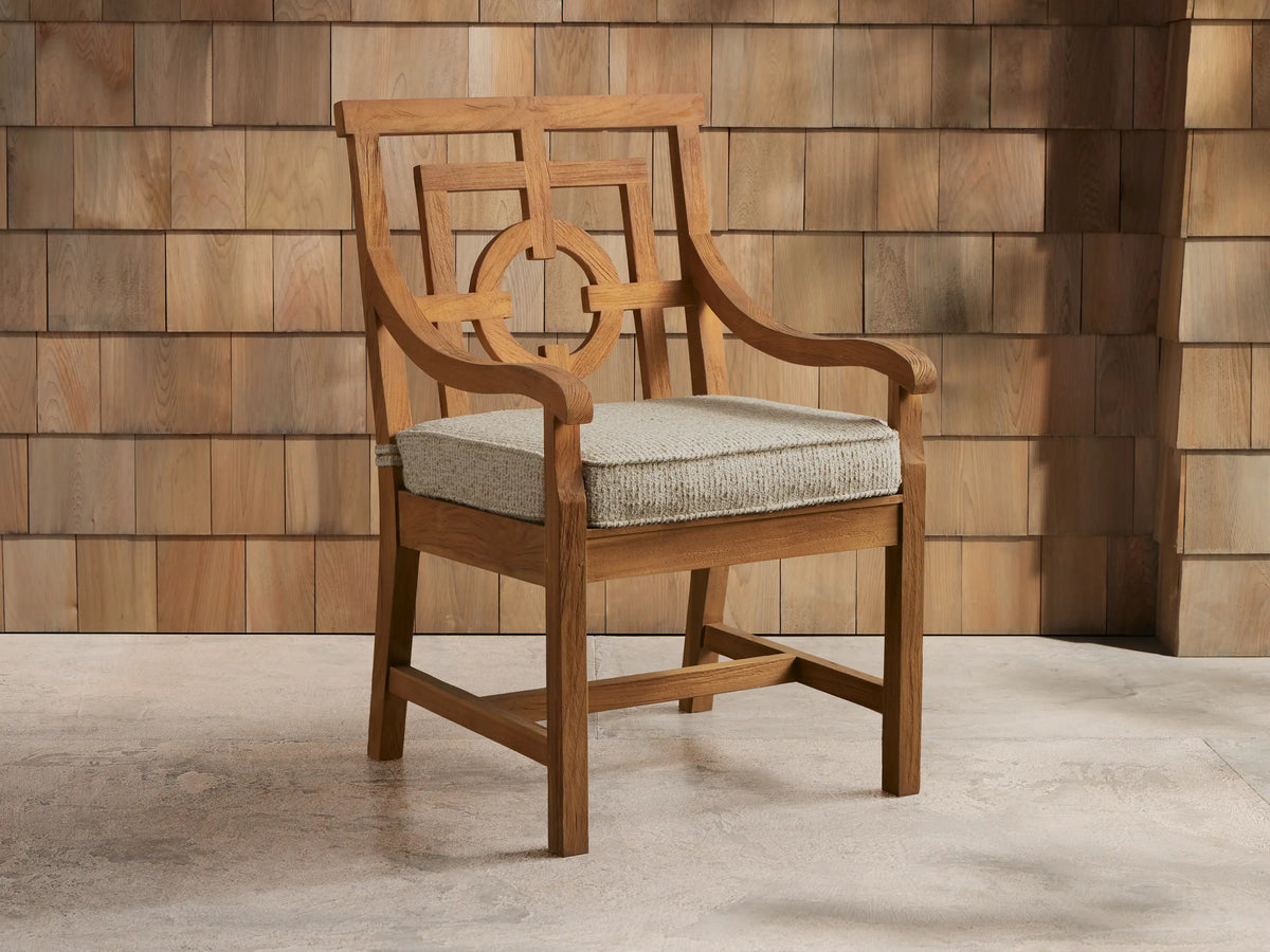 Hamptons Outdoor Dining Arm Chair, teak wood, light grey cushion, intricate back, on patio against wood shingle wall.