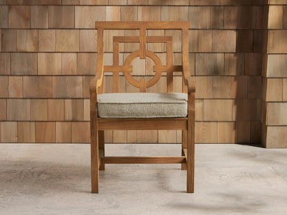 Hamptons Outdoor Dining Arm Chair, teak wood, light grey cushion, intricate back, on patio against wood shingle wall.