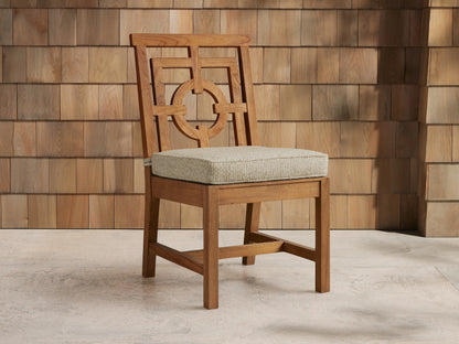 Hamptons Outdoor Dining Side Chair, natural teak wood, ornate geometric backrest, light beige cushion, on a stone patio with a wood shingle wall.