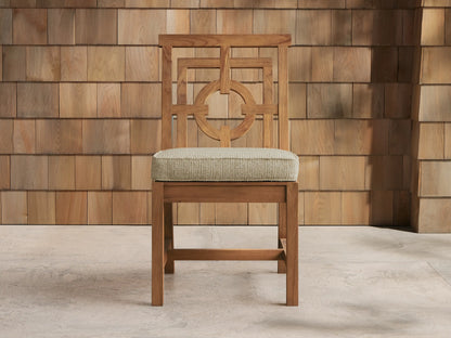Hamptons Outdoor Dining Side Chair, natural teak wood, ornate geometric backrest, light beige cushion, on a stone patio with a wood shingle wall.
