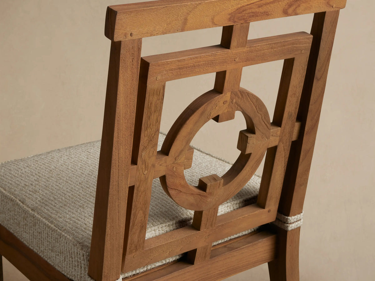 Hamptons Outdoor Dining Side Chair, natural teak with geometric backrest, beige cushion, against a light wood shingle wall.