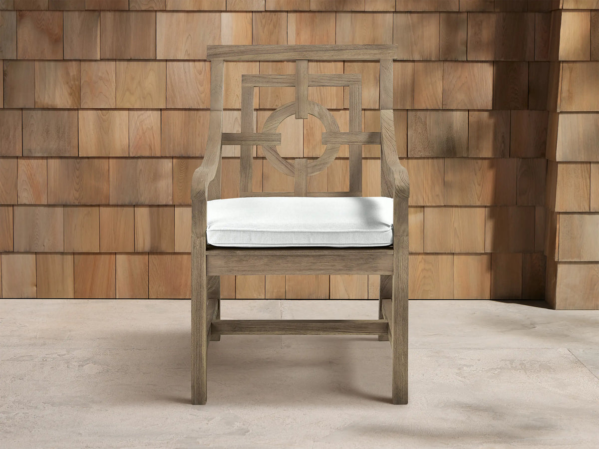 Hamptons Outdoor Dining Arm Chair in weathered teak, white cushion, intricate geometric backrest. Front view against shingle wall.