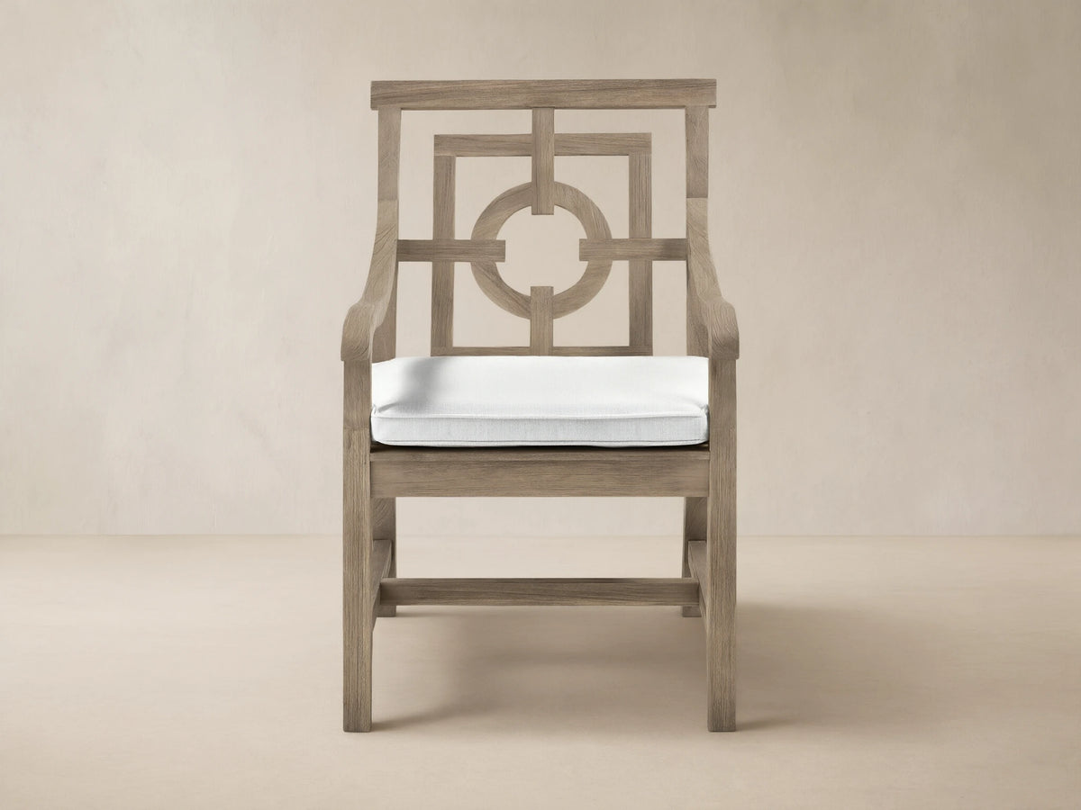 Hamptons Outdoor Dining Arm Chair in weathered teak, white cushion, intricate geometric backrest. Front view against shingle wall.